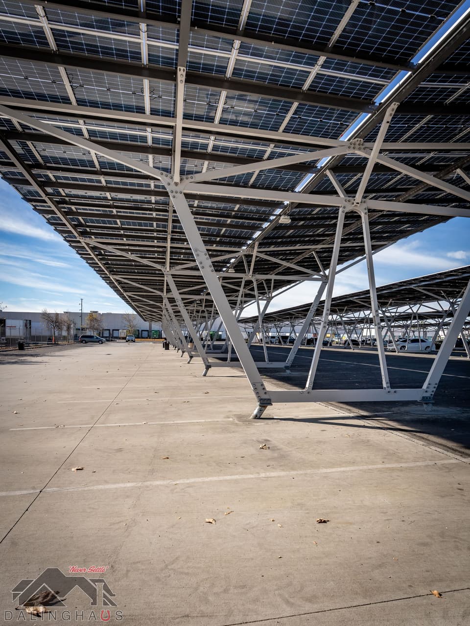 View from beneath the canopy along a line of white steel columns, sunlight coming through the gaps in the panel deck onto an empty concrete parking bay.