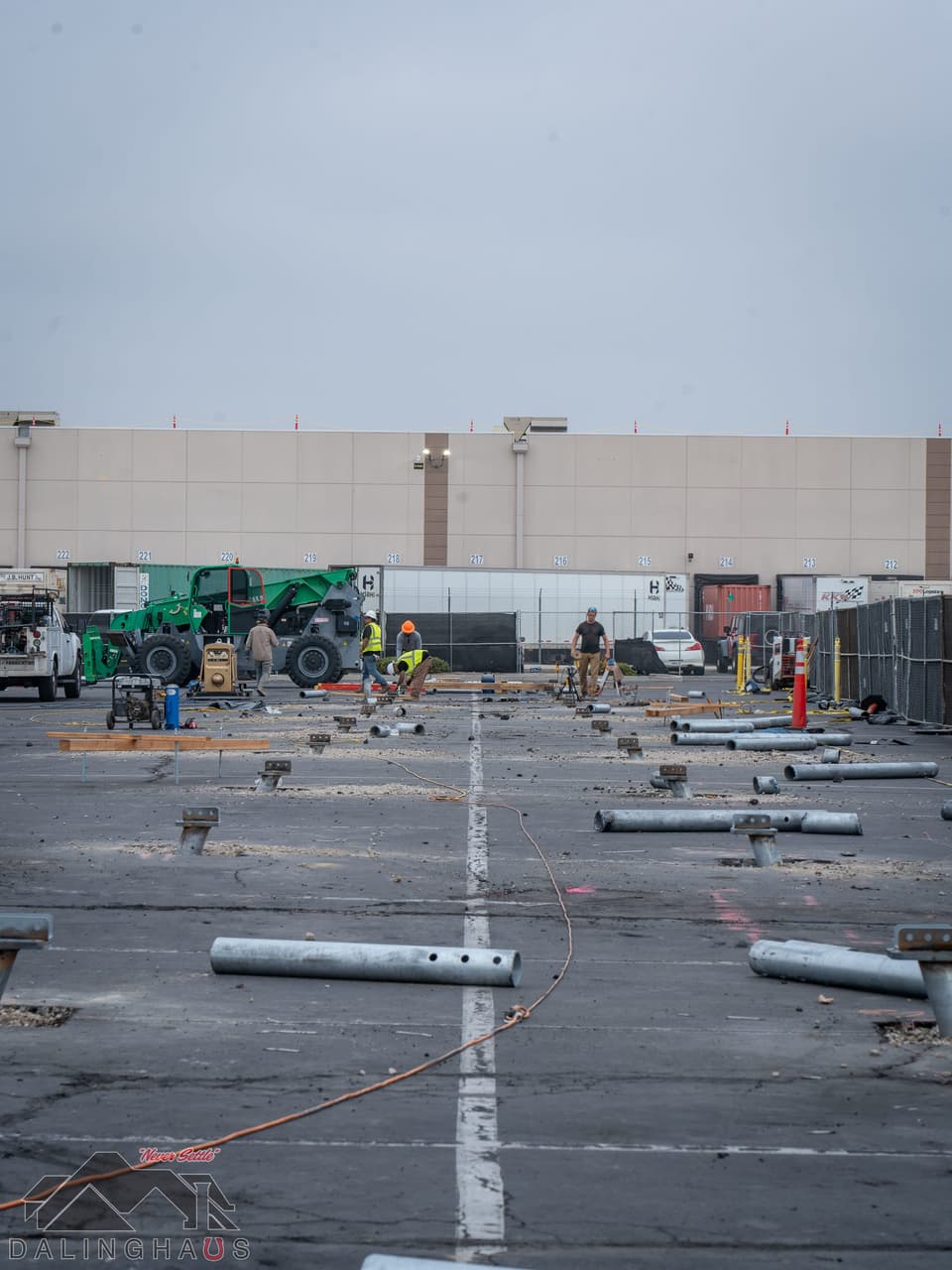 Wide view across an asphalt parking lot with galvanized helical pile shafts laid out in rows on the pavement, crew and green equipment working along a long tan warehouse wall under an overcast sky.