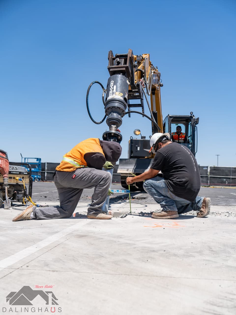 Two crew members kneeling at ground level at the base of a tracked excavator fitted with a hydraulic drive head, working at the pile location under a clear blue sky.