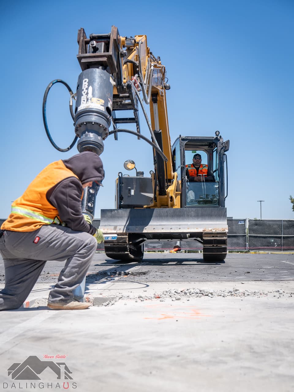 Hydraulic torque drive head raised on the boom of a tracked excavator, a crew member crouched at the pile location below, operator visible in the cab, against a deep blue sky.