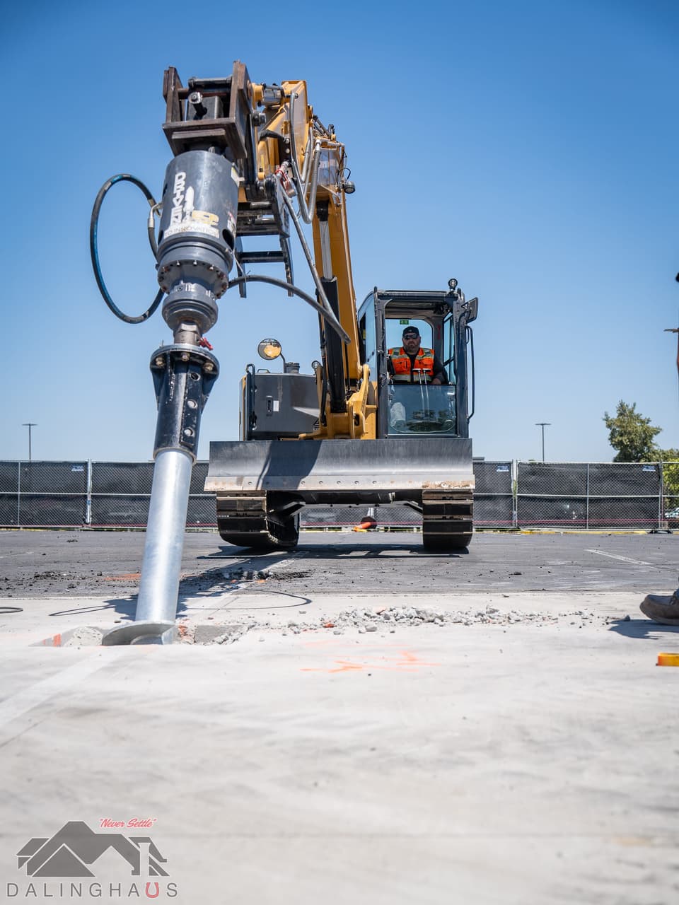 Tracked CAT excavator with a hydraulic torque head turning a galvanized helical pile shaft into an opening cut in a concrete parking lot, broken concrete around the hole and orange utility marks sprayed on the deck.