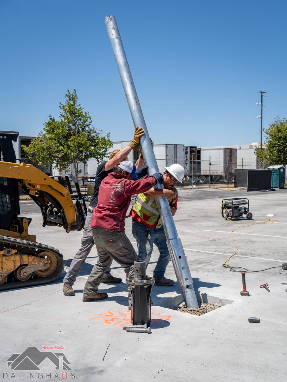 Crew guiding a galvanized pile shaft into position over a cut opening in a concrete lot, a black pile cap fixture standing on the deck in the foreground and a skid steer at the left.