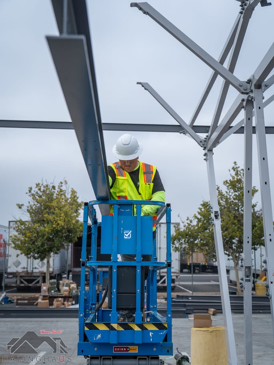 Worker in a white hard hat and high visibility vest standing in a blue vertical mast lift, setting a gray steel arm into the branching head of a white canopy column, with trailers and trees behind under an overcast sky.