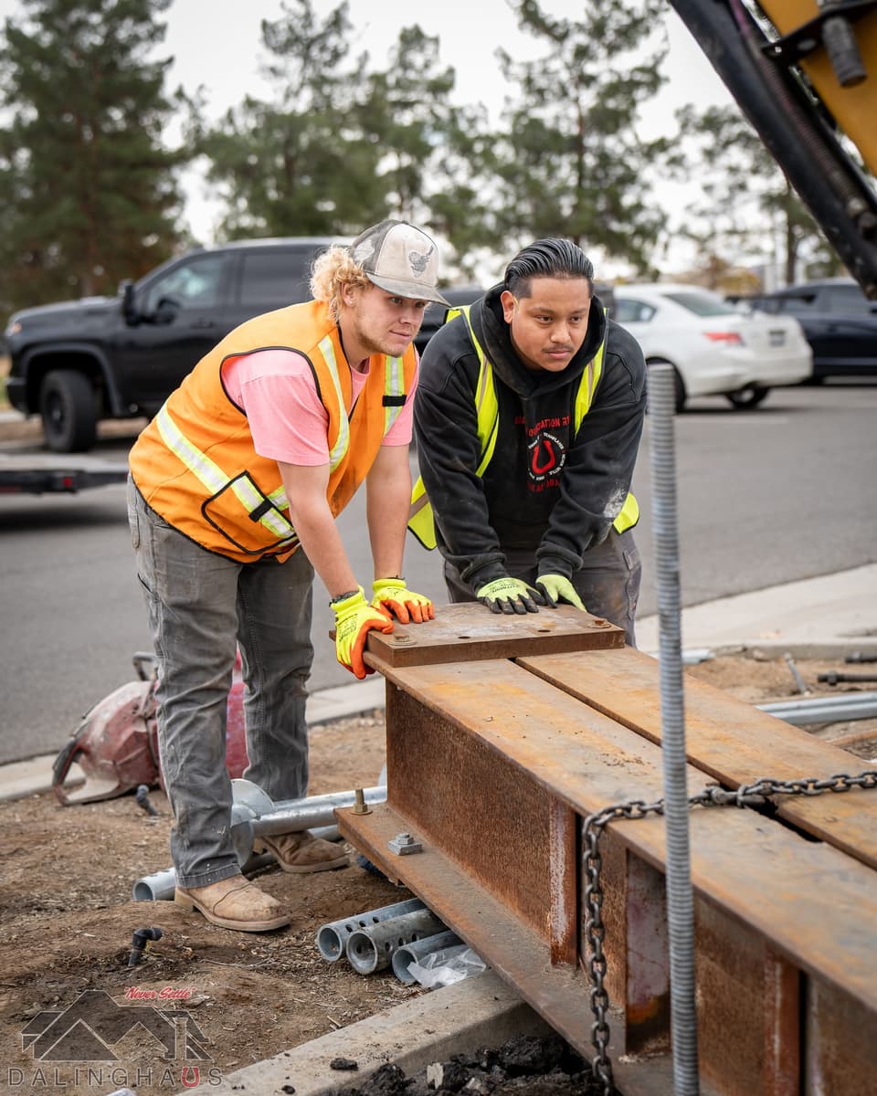 Two crew members in high visibility vests positioning a heavy rusted steel reaction beam over a test pile, threaded rod and chain rigged at the pile head.