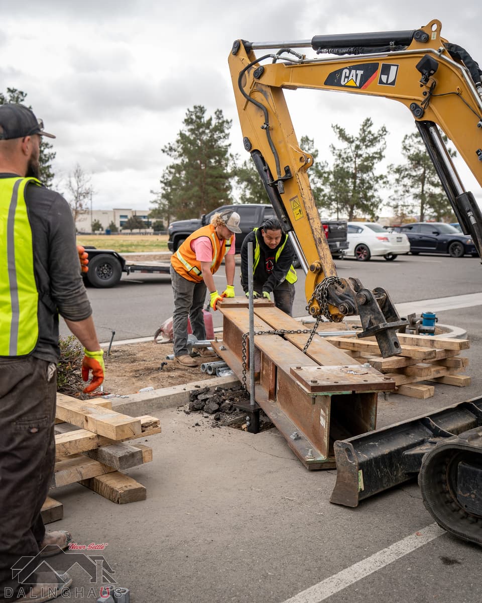 Crew rigging a rusted steel reaction beam on chains from the boom of a CAT mini excavator, with timber cribbing stacked alongside on the asphalt.