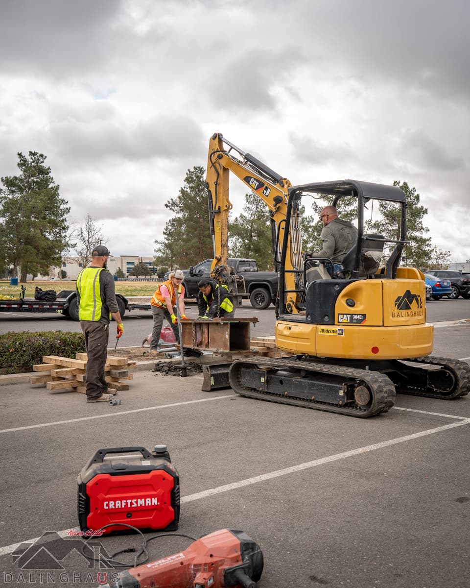 Pull test underway in a parking lot: a Dalinghaus branded CAT 304E2 mini excavator with an operator, three crew working around a rusted steel reaction beam set on timber cribbing over a test pile, on an overcast day.