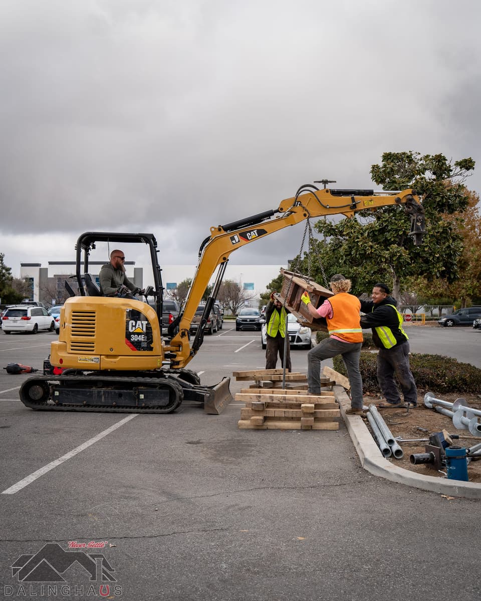 Crew and a CAT mini excavator setting timber cribbing and a steel reaction beam beside a landscaped parking island, with a road and low buildings behind.