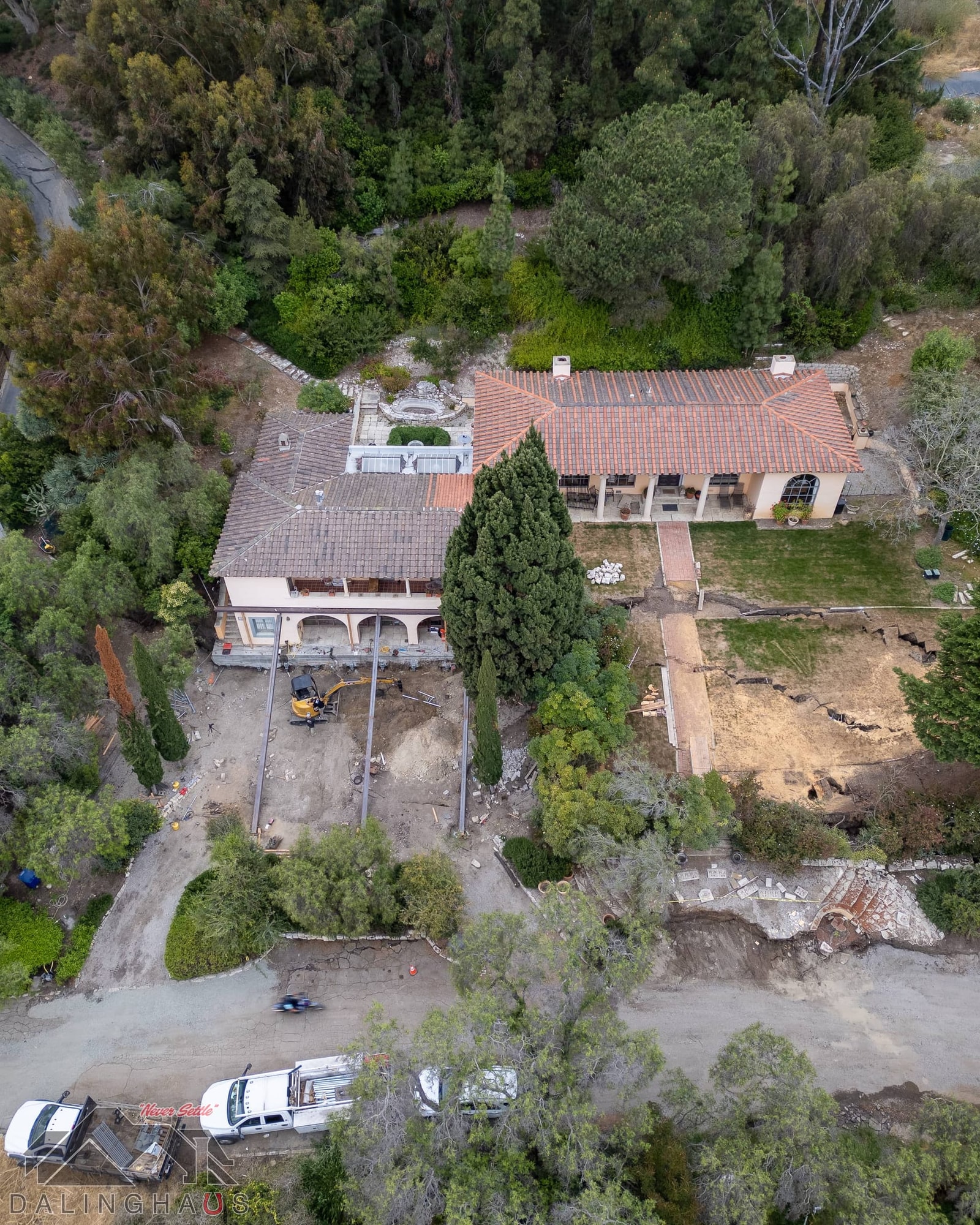 Aerial view of a tile roof home, steel beams and a mini excavator under one wing, lawn split by fissures