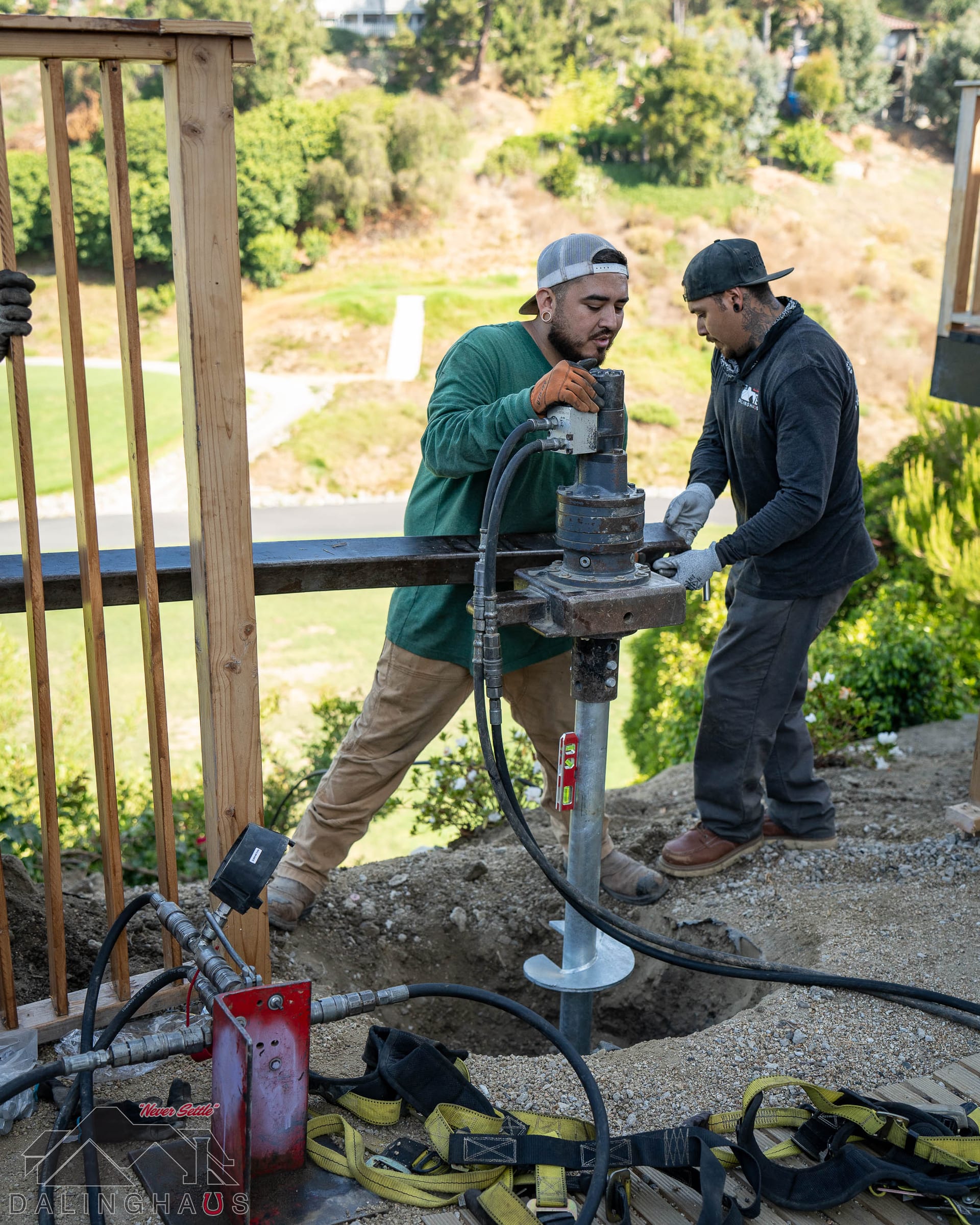 Two crew on a hydraulic drive head, helix plate in the pit, deck railing and green hillside behind