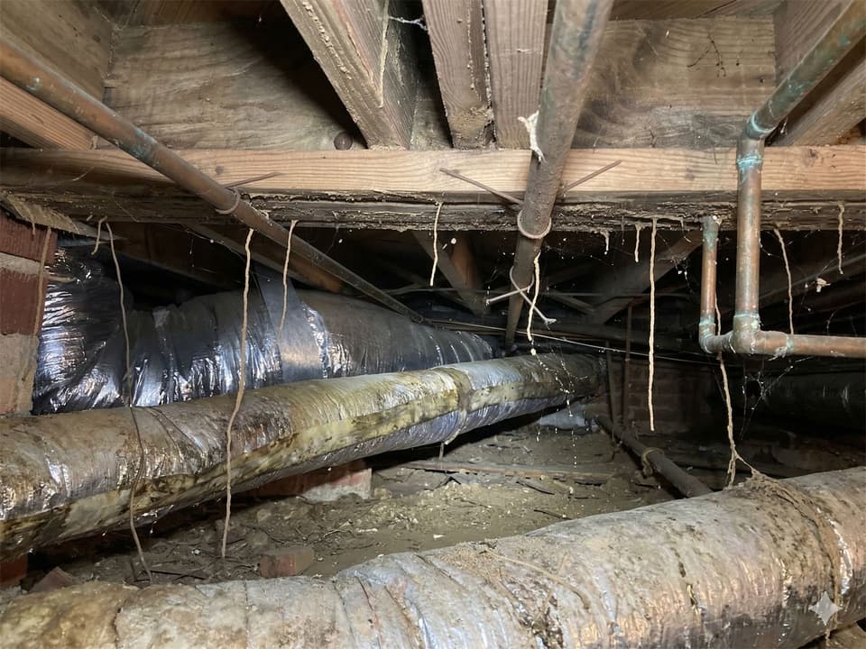 A damp crawl space with sagging ductwork, old copper pipes and debris on the soil