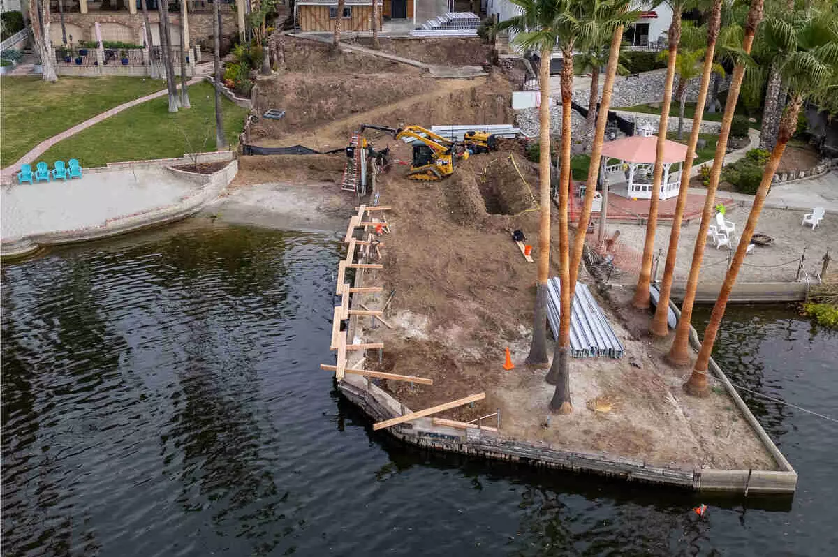 Aerial view of a lakefront seawall rebuild, with timber shoring along the water, an excavator and a row of palm trees on the lot