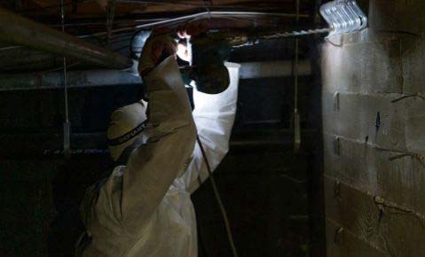 Worker in a protective suit drilling into a concrete block stem wall in a dark crawl space