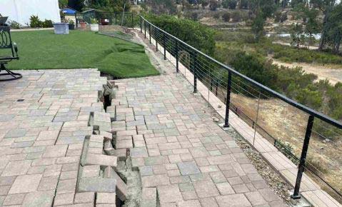 Paver patio split and buckled along a crack beside a railing at the edge of a slope