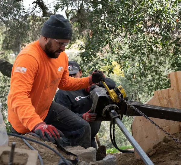 Crew member in safety glasses and gloves guiding a hydraulic drive head as it turns a steel shaft into a hillside