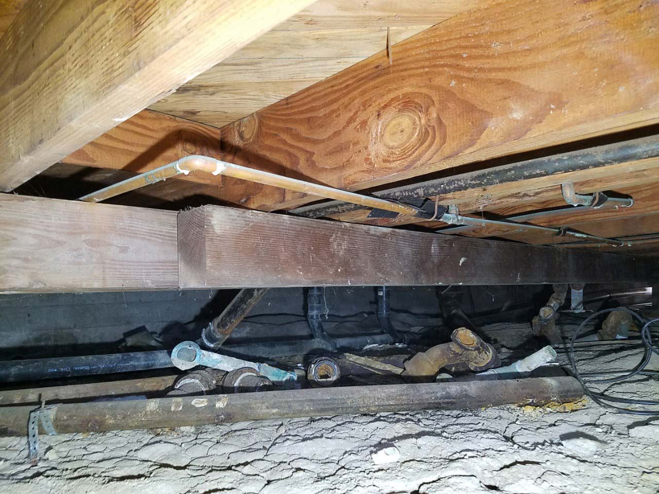 Wooden girder and floor framing seen from inside a crawl space, with pipes running beneath
